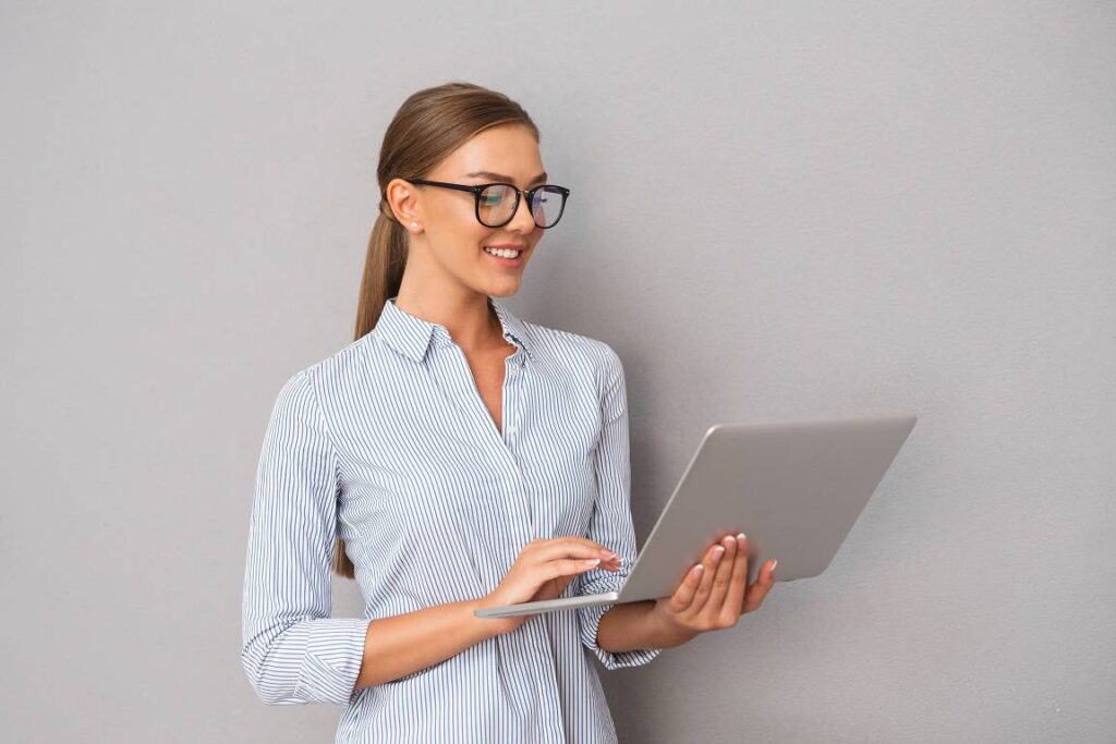 Imagen de hermosa mujer de negocios sonriente de pie sobre fondo de pared gris usando ordenador portátil. Venta de inmuebles, análisis de mercado